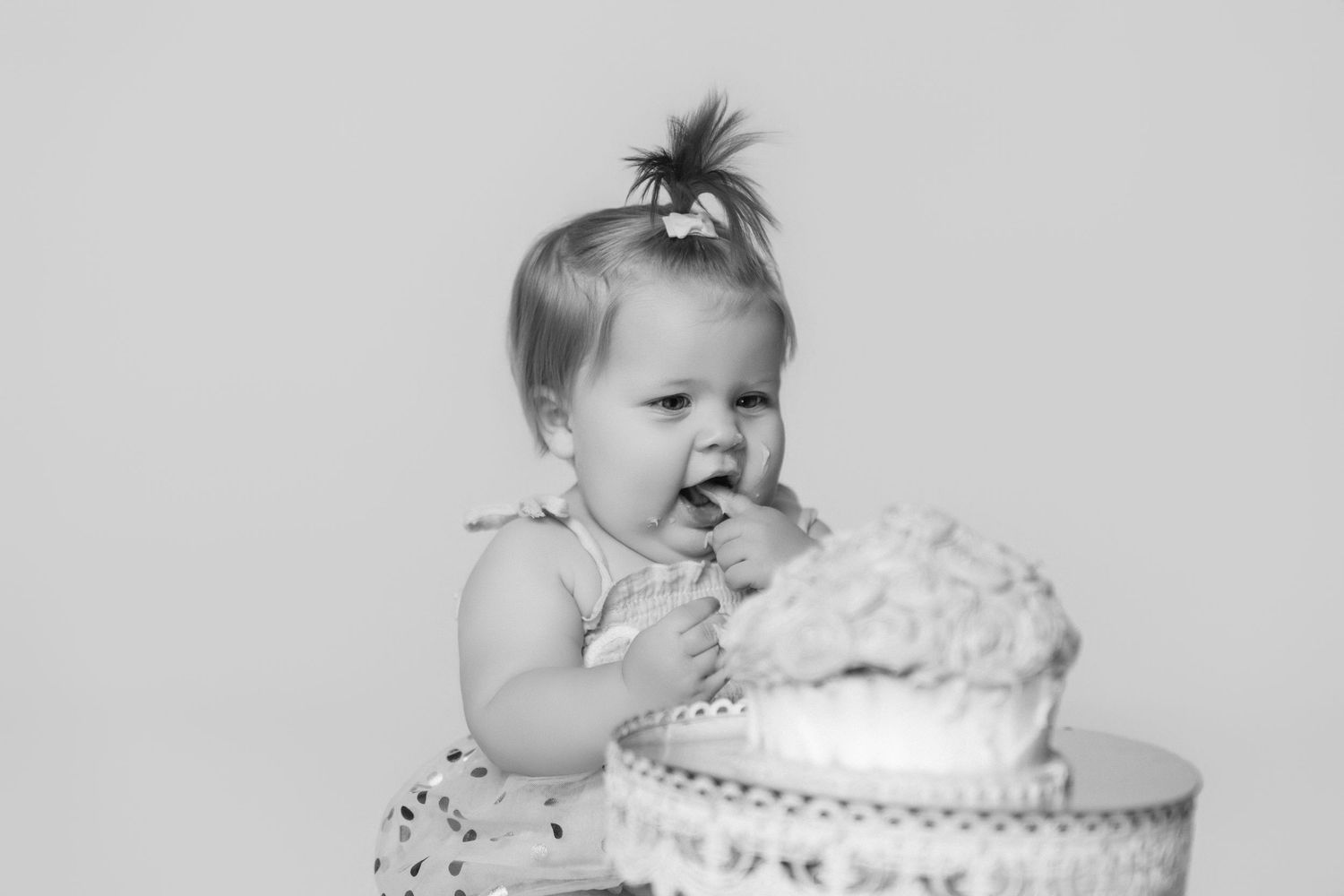 Black and white photo of baby reaching for cake on pedestal during cake smash session.
