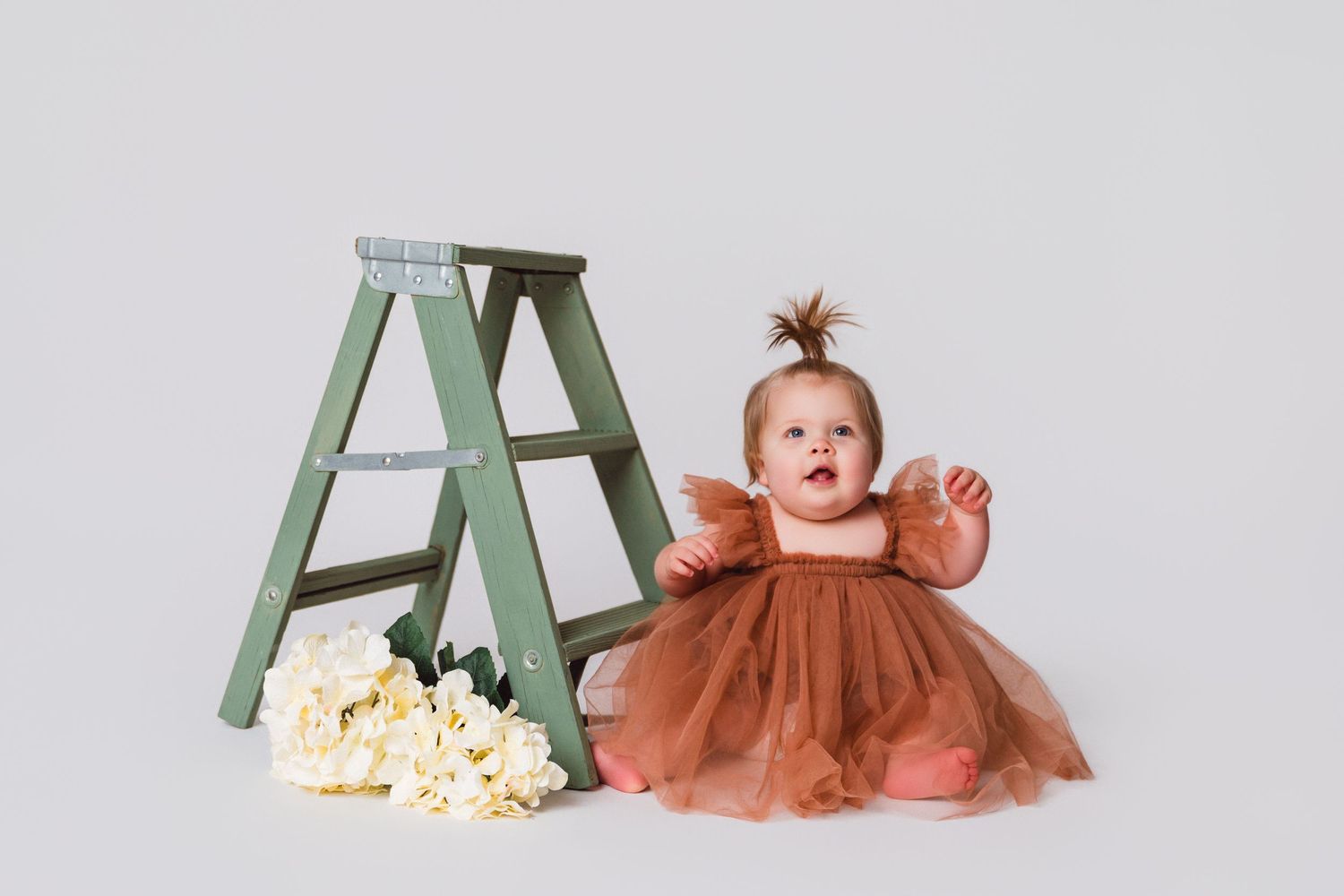 Baby in rust-colored tulle dress sitting by mint green ladder with flowers.