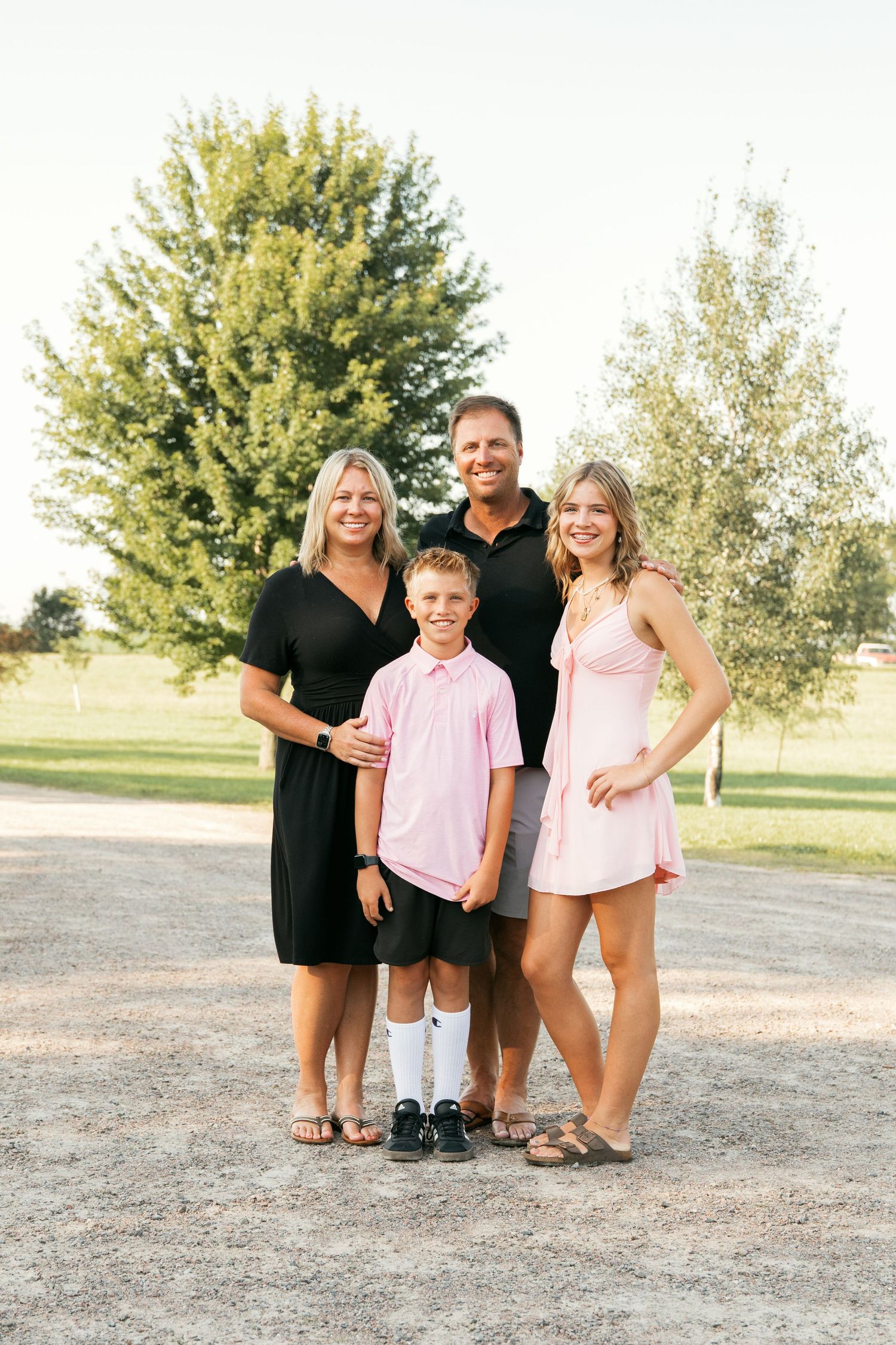 A family wearing coordinated black and pink outfits poses together outdoors on a dirt path near trees.