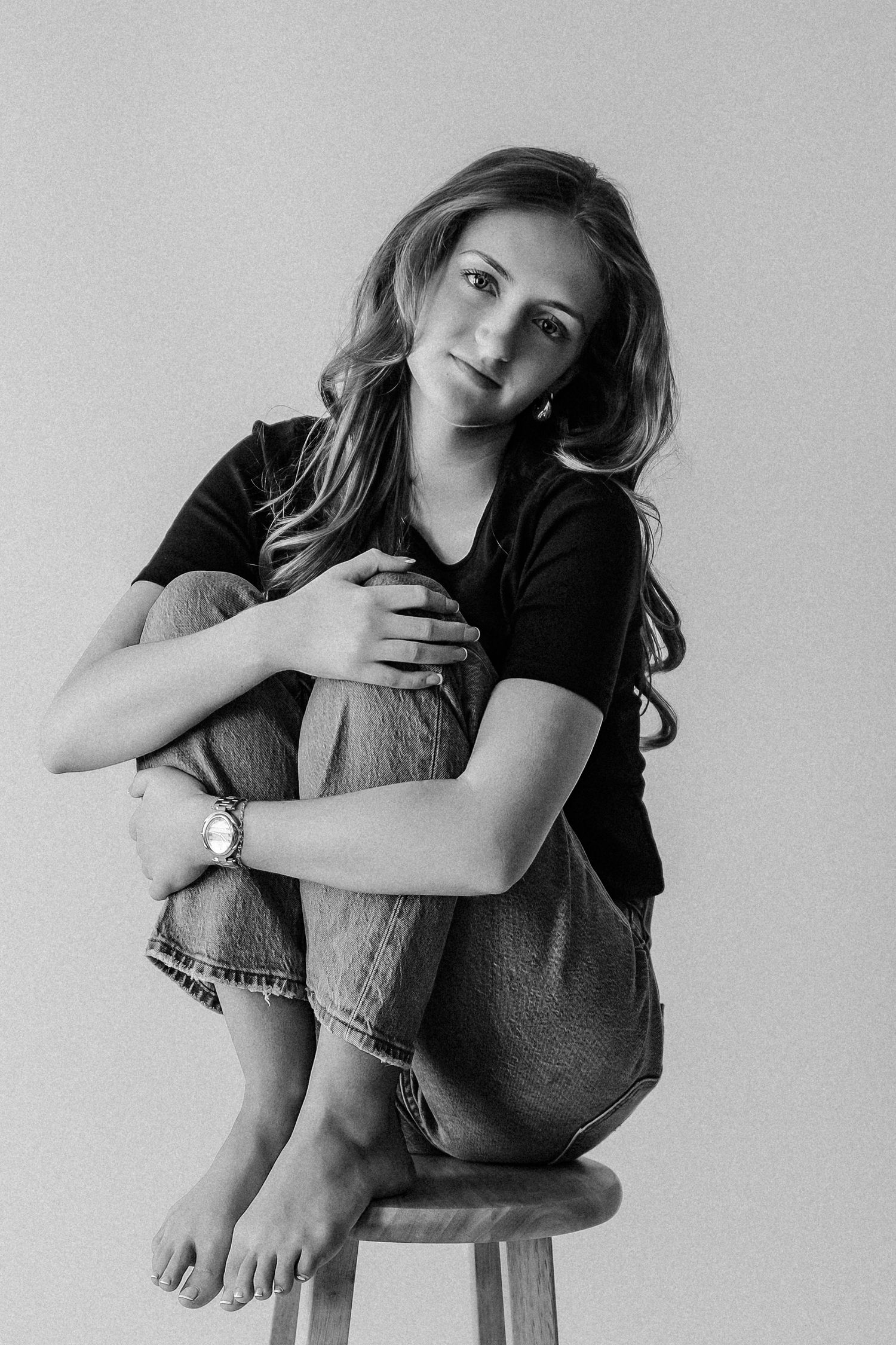 Black and white photo of young woman hugging knees on wooden stool, wearing black tee and jeans, barefoot.