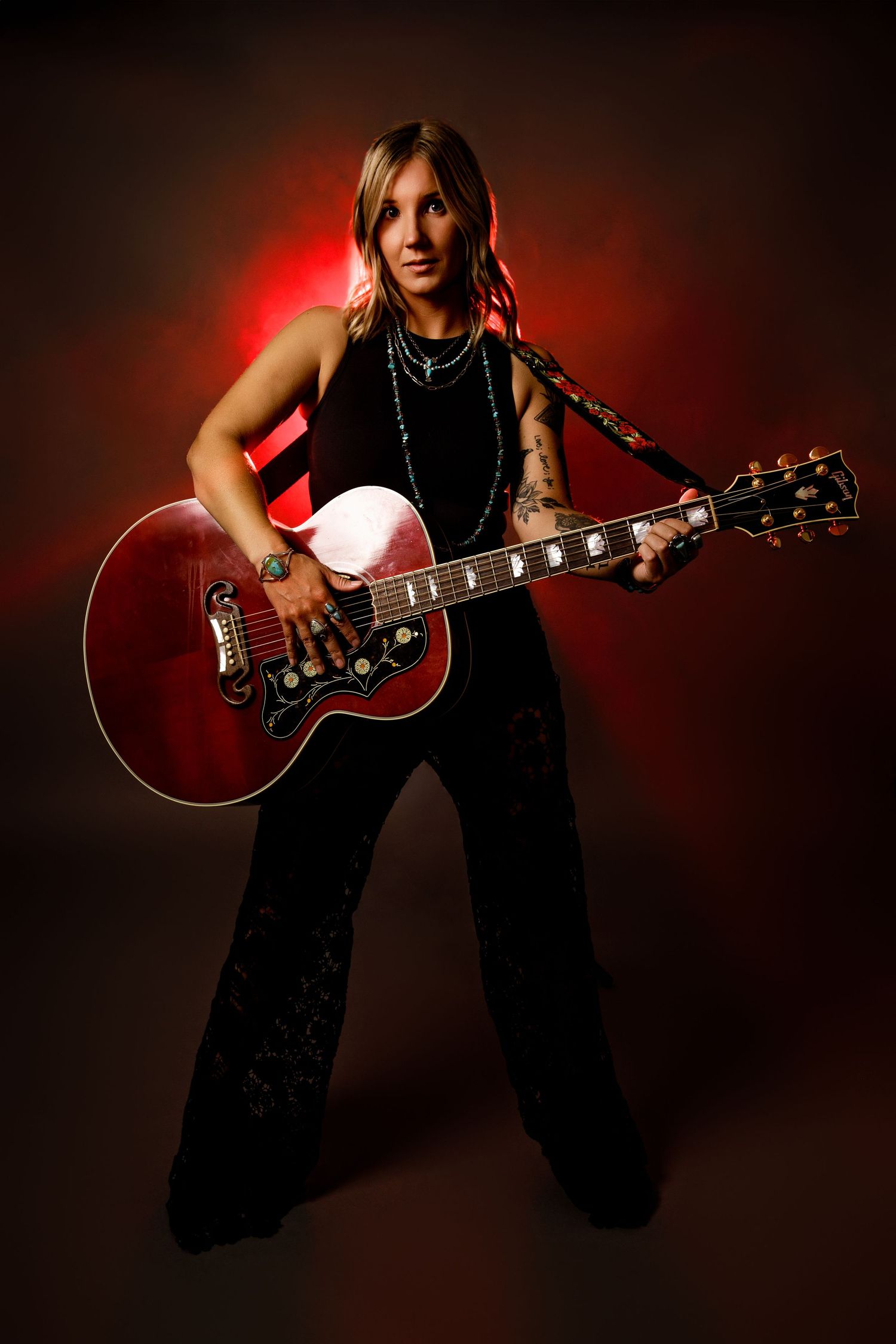 Female guitarist playing red Gibson acoustic guitar under dramatic red lighting in black outfit.