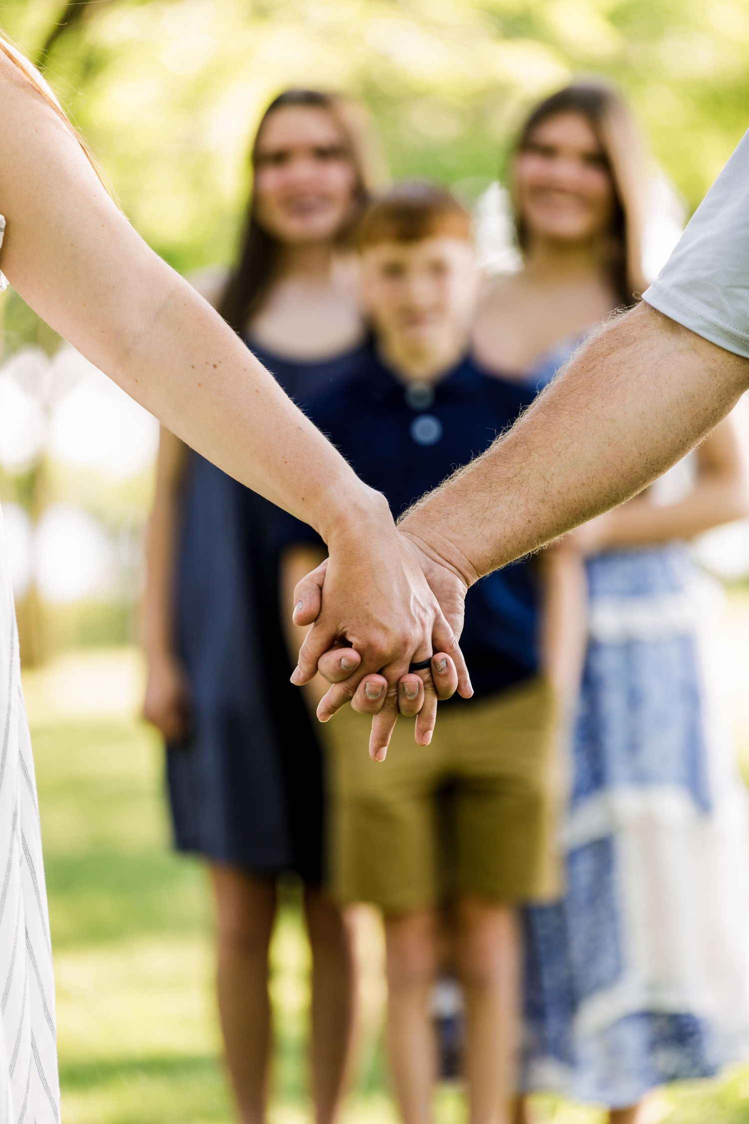 Couple holding hands outdoors with three children smiling in the background in a lush green park.