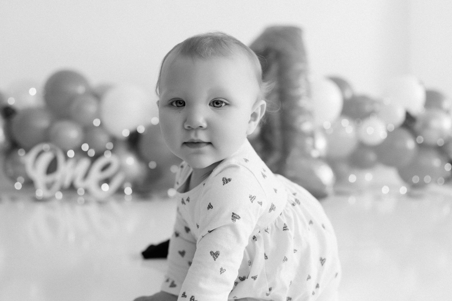Black and white portrait of baby sitting on floor with party decorations in background.
