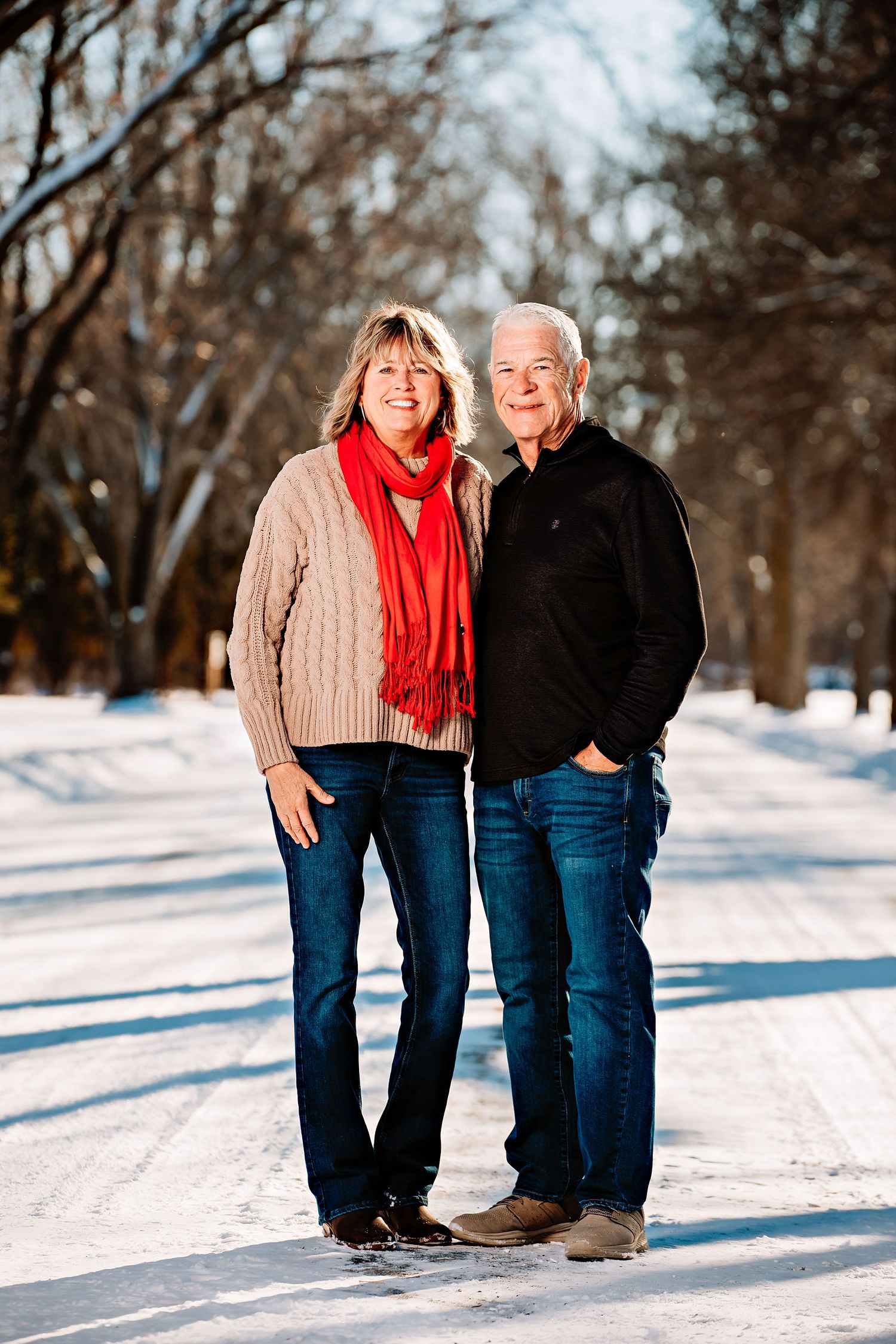 A couple stands together on a snow-covered path wearing winter attire and denim.