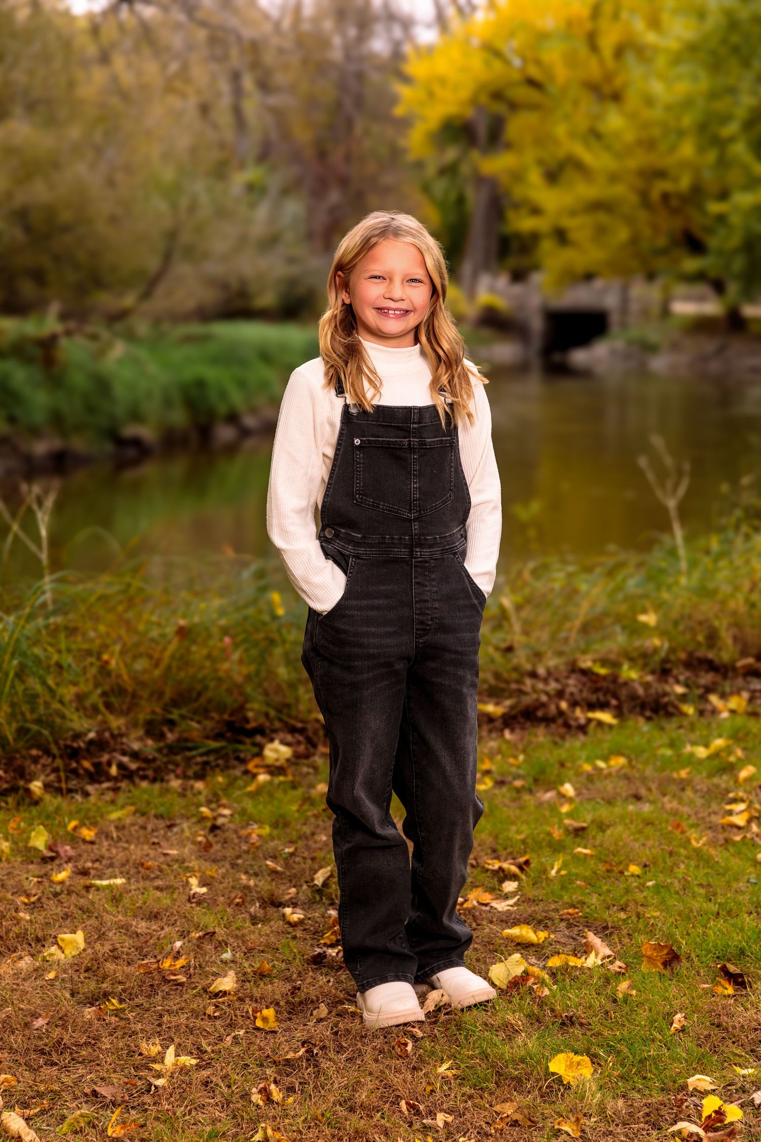 Child wearing black overalls and white shirt plays in autumn leaves by a lake.