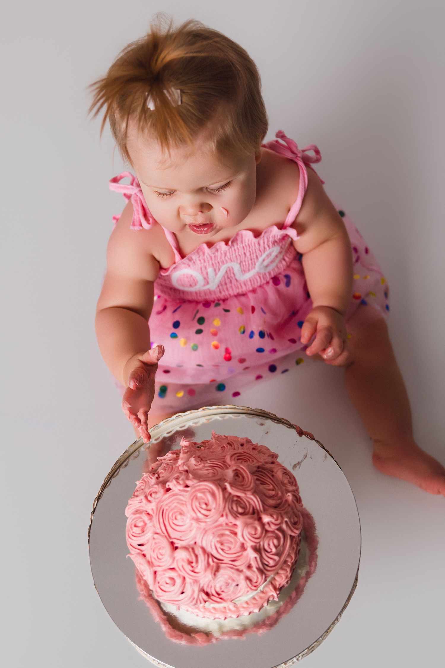 Overhead view of baby in pink dress looking down at rose-decorated pink birthday cake.