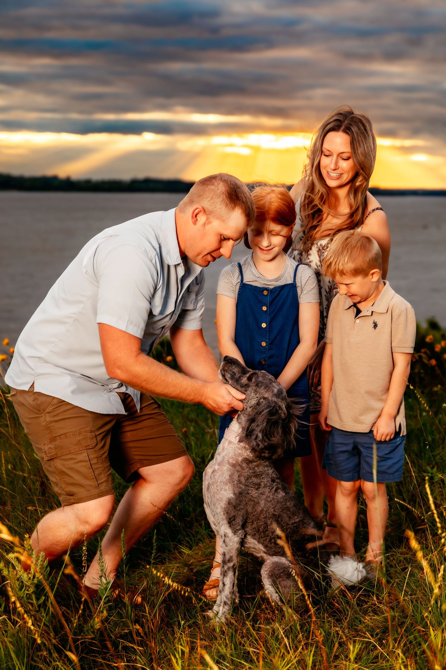 A group shares a warm sunset moment with their dog in a grassy field by the water.
