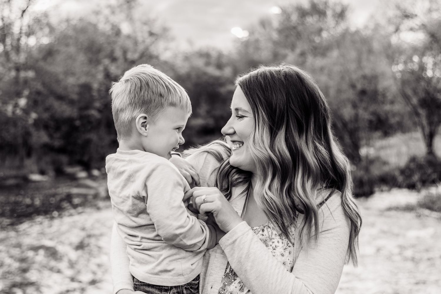 Black and white photo of a smiling mother holding her young toddler son outdoors among trees.