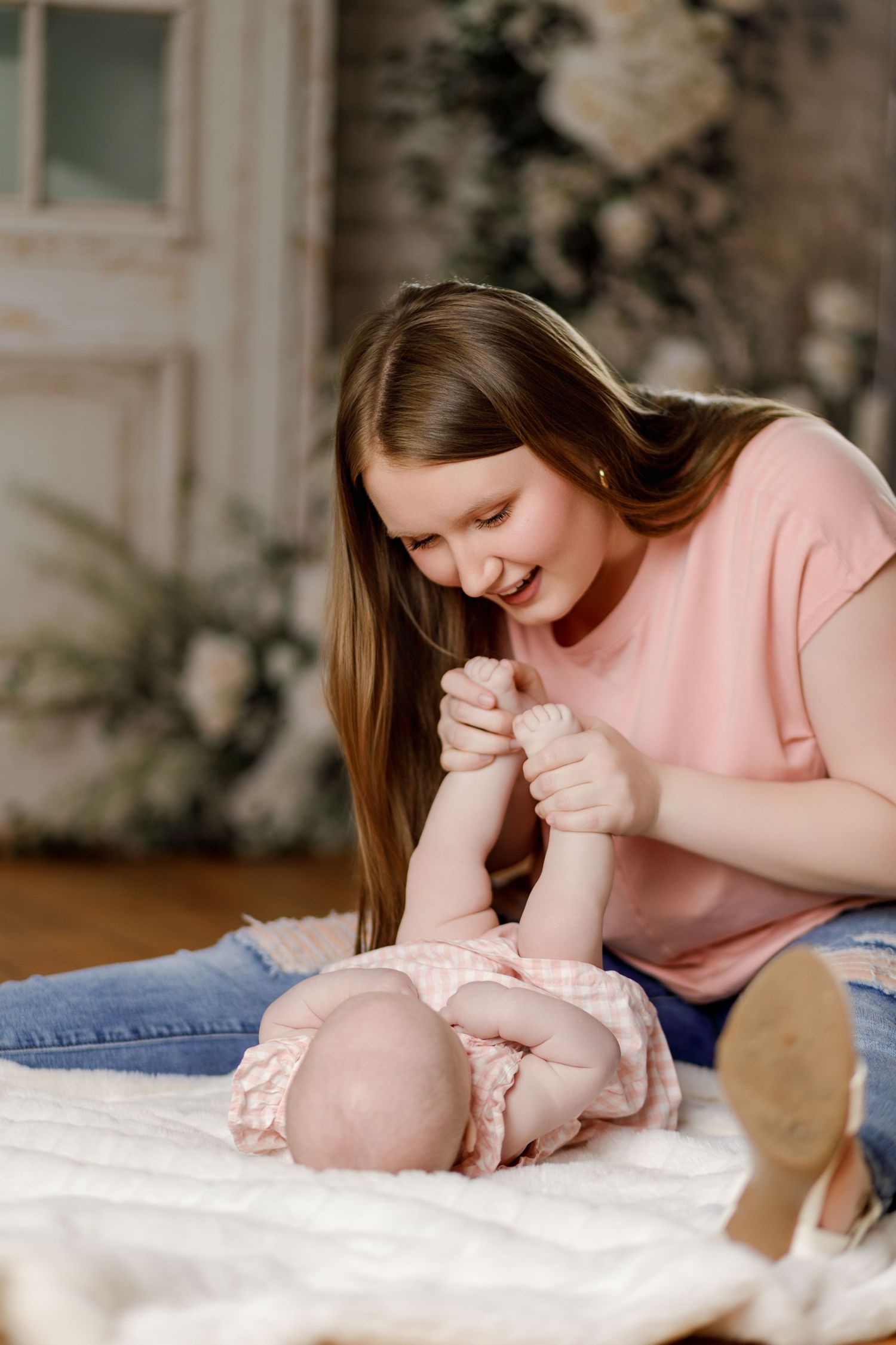 A smiling young mother in a pink shirt and jeans plays with her newborn baby lying on a white blanket.