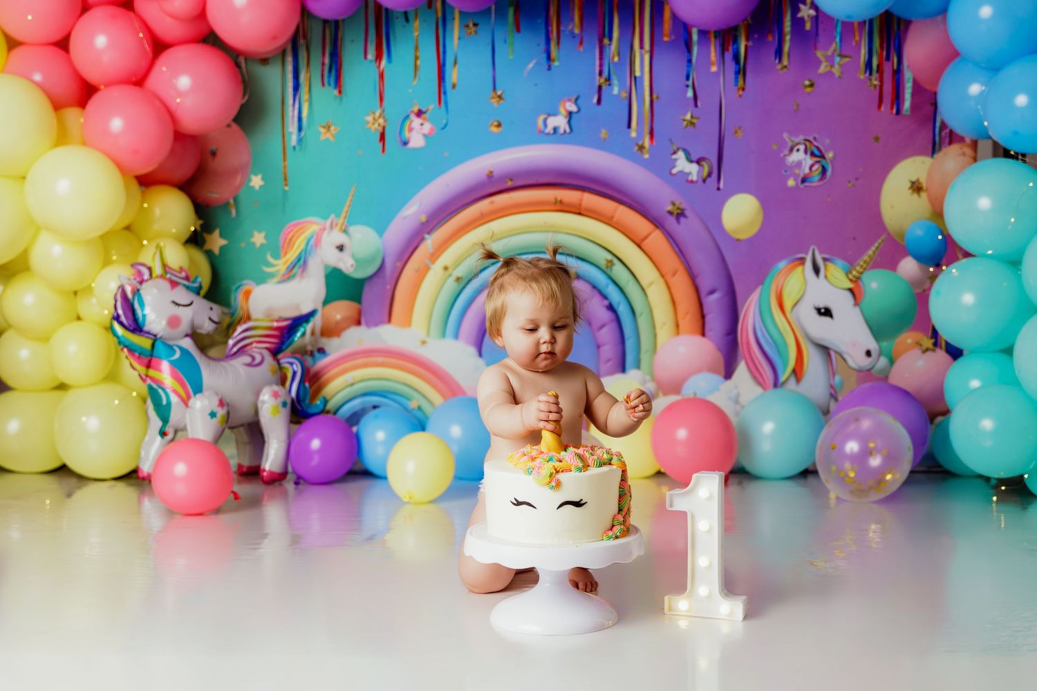 Baby girl enjoying unicorn first birthday cake smash with colorful rainbow balloons and festive backdrop.