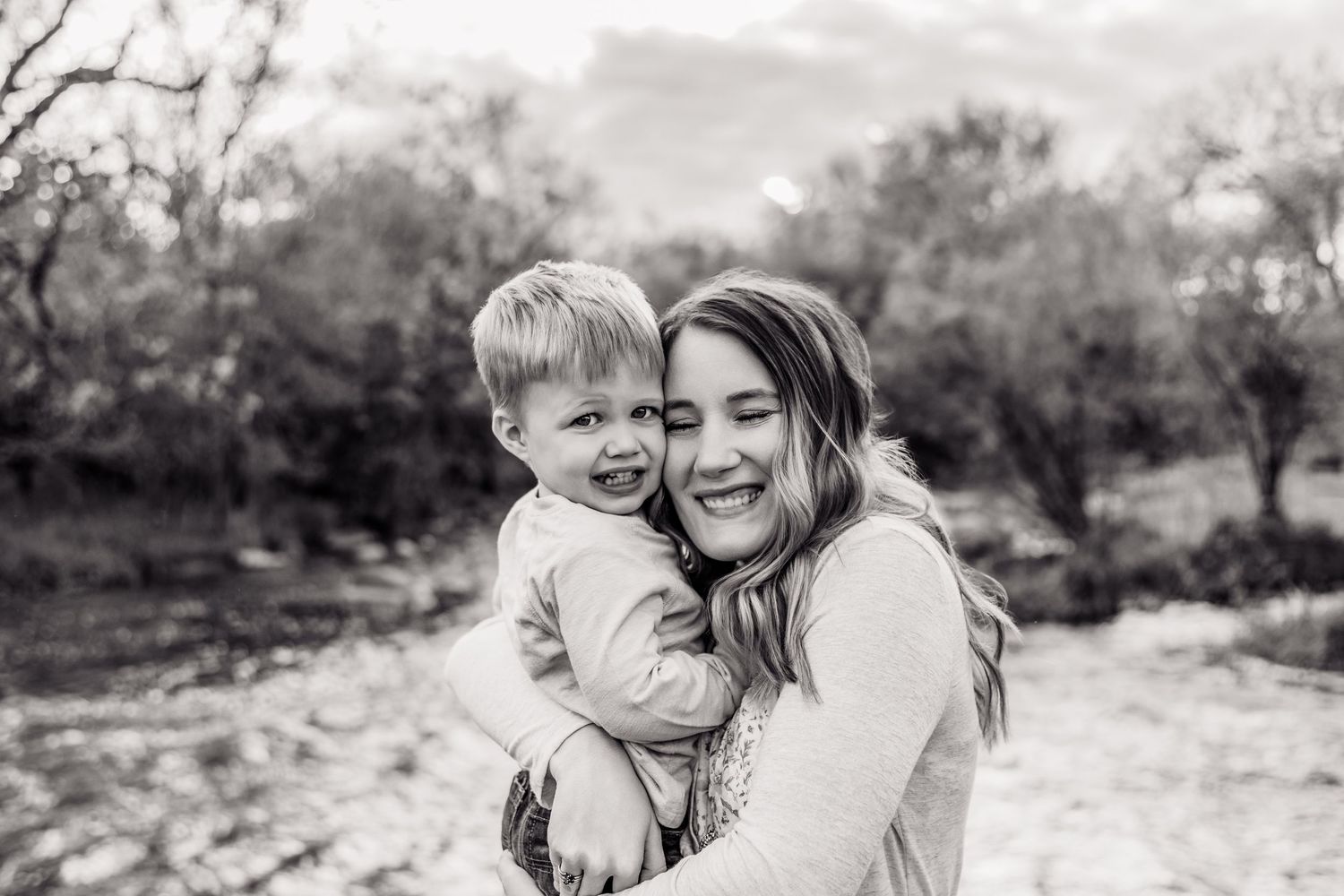Black and white photo of a laughing woman holding a smiling toddler outdoors among trees.