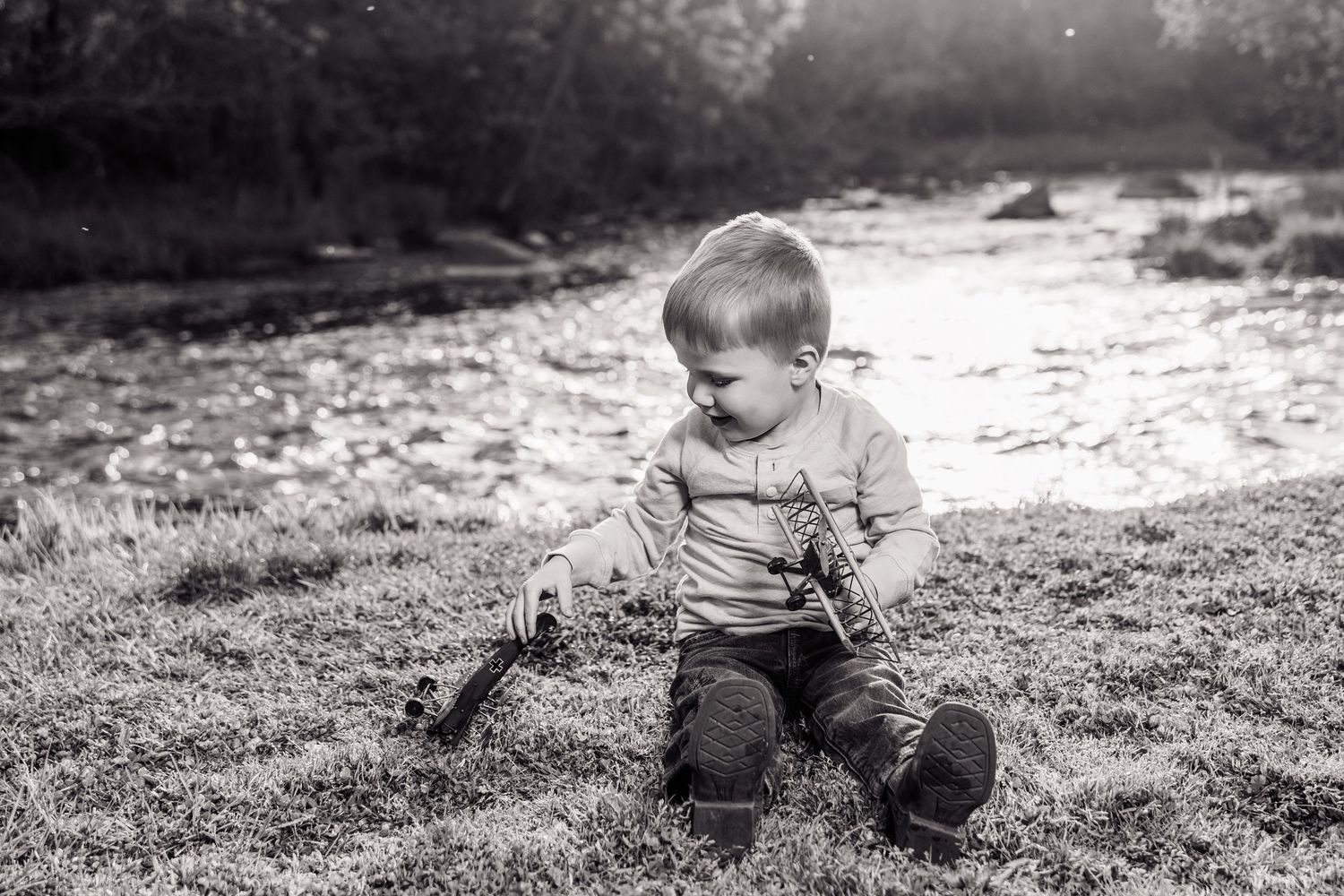Black and white photo of a toddler boy sitting on grass near a river, playing with a toy outdoors.