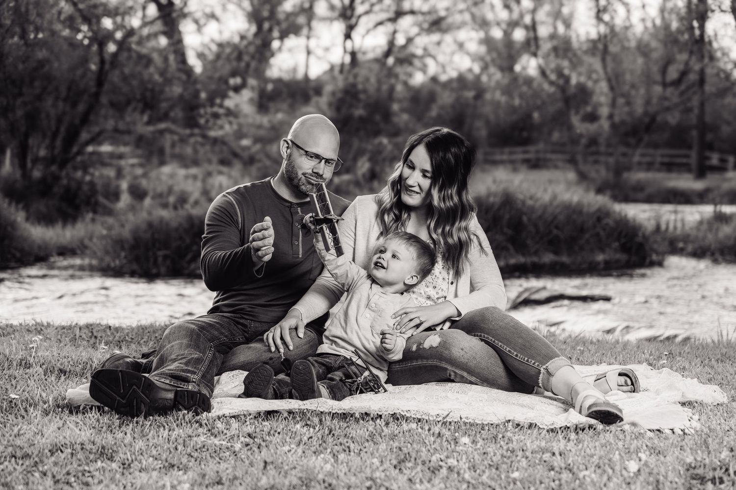 Black and white family photo of parents and baby sitting on blanket in park, enjoying a peaceful outdoor moment together.