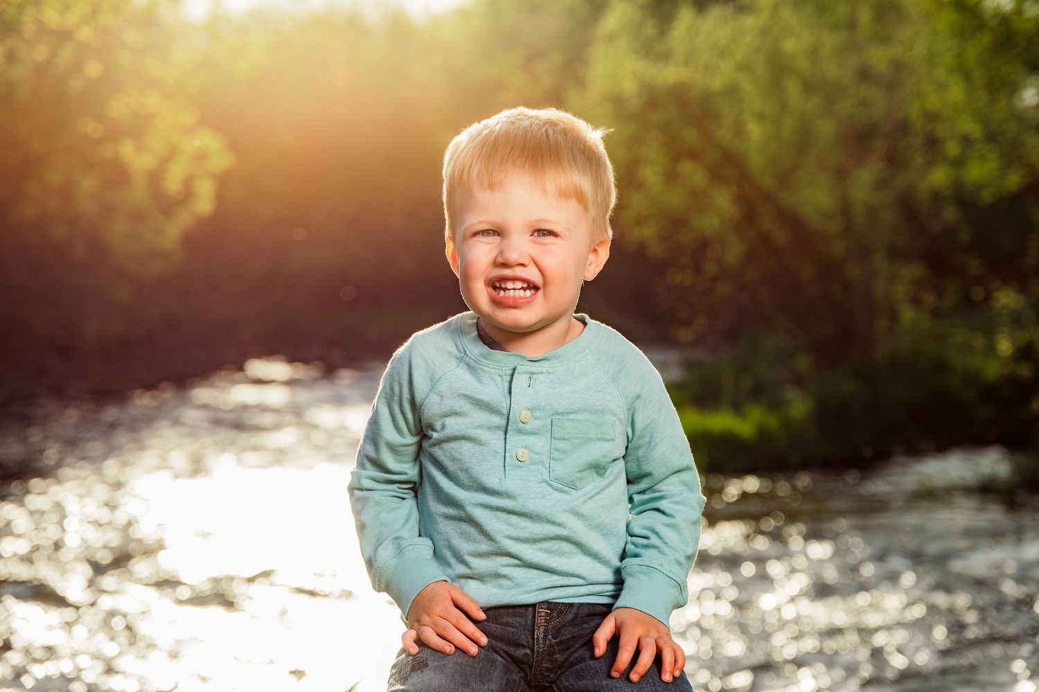 Cheerful toddler boy with blonde hair smiling outdoors near a sunlit river in a teal long-sleeve shirt and jeans.