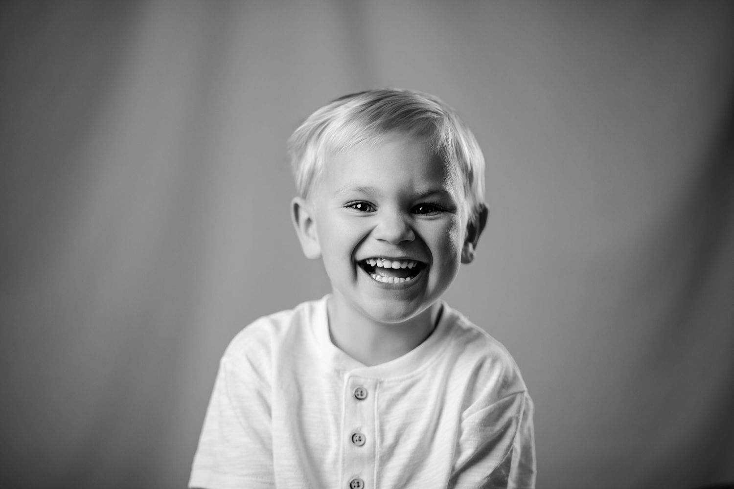 Joyful blond boy in white henley shirt laughing in black and white studio portrait photo.