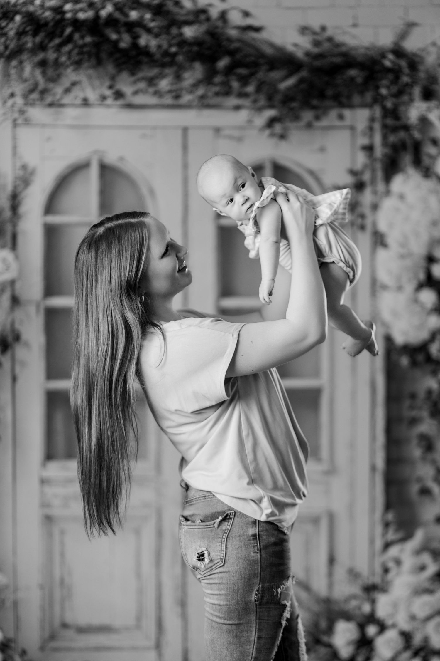 Black and white photo of a mother lifting her baby overhead in a flower-decorated studio.