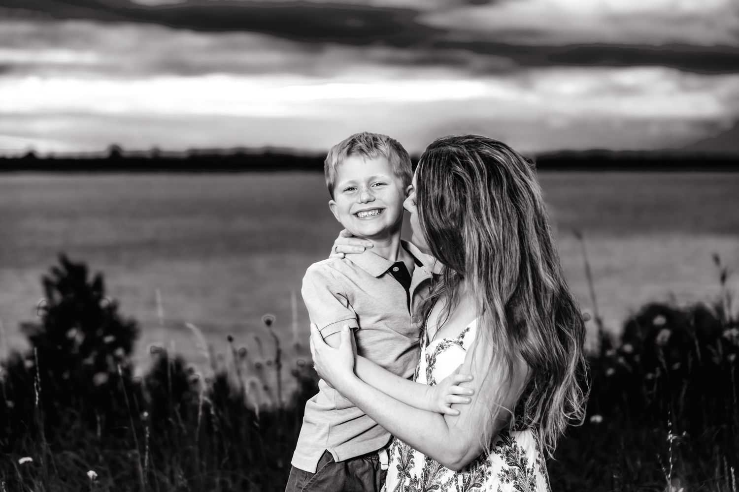 Black and white portrait of a tender moment between parent and child outdoors against a dramatic cloudy sky.