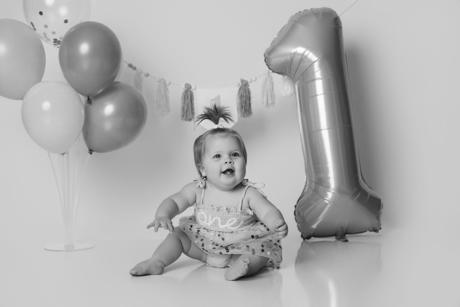Black and white photo of baby with balloons and silver number one birthday decoration.