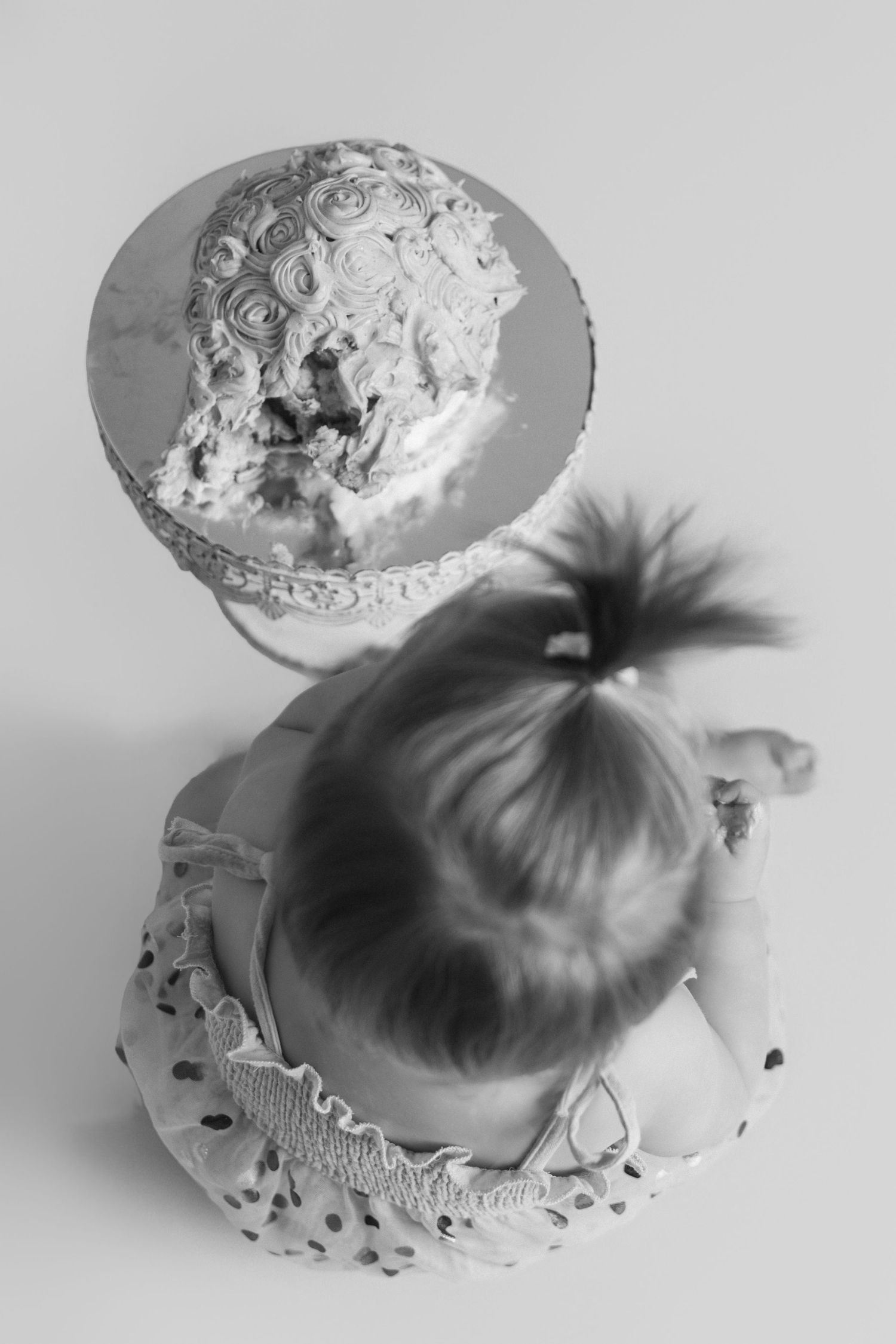 Black and white aerial view of baby examining rose-decorated cake on round platter.