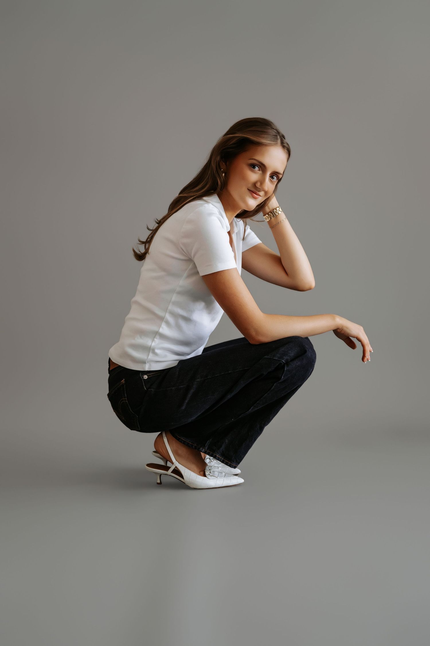 Woman in white tee and dark jeans crouching and smiling, white heels, studio portrait.
