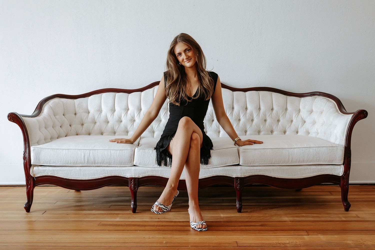 Smiling young woman in black dress and animal print heels sitting on ornate white tufted sofa on hardwood floor.