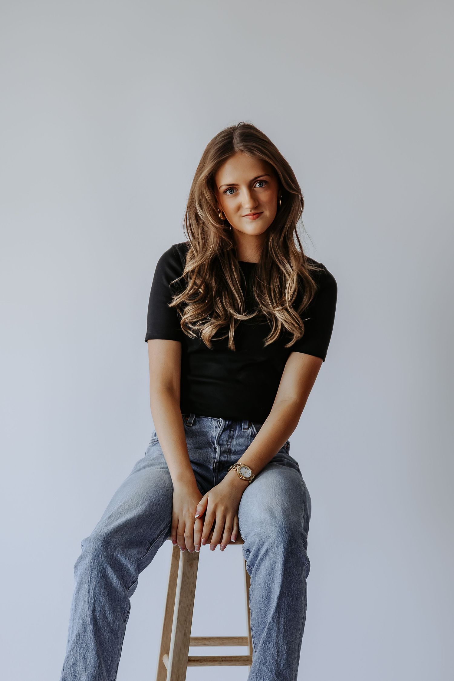 Young woman with wavy hair in black top and gray jeans seated on wooden stool in minimalist studio.