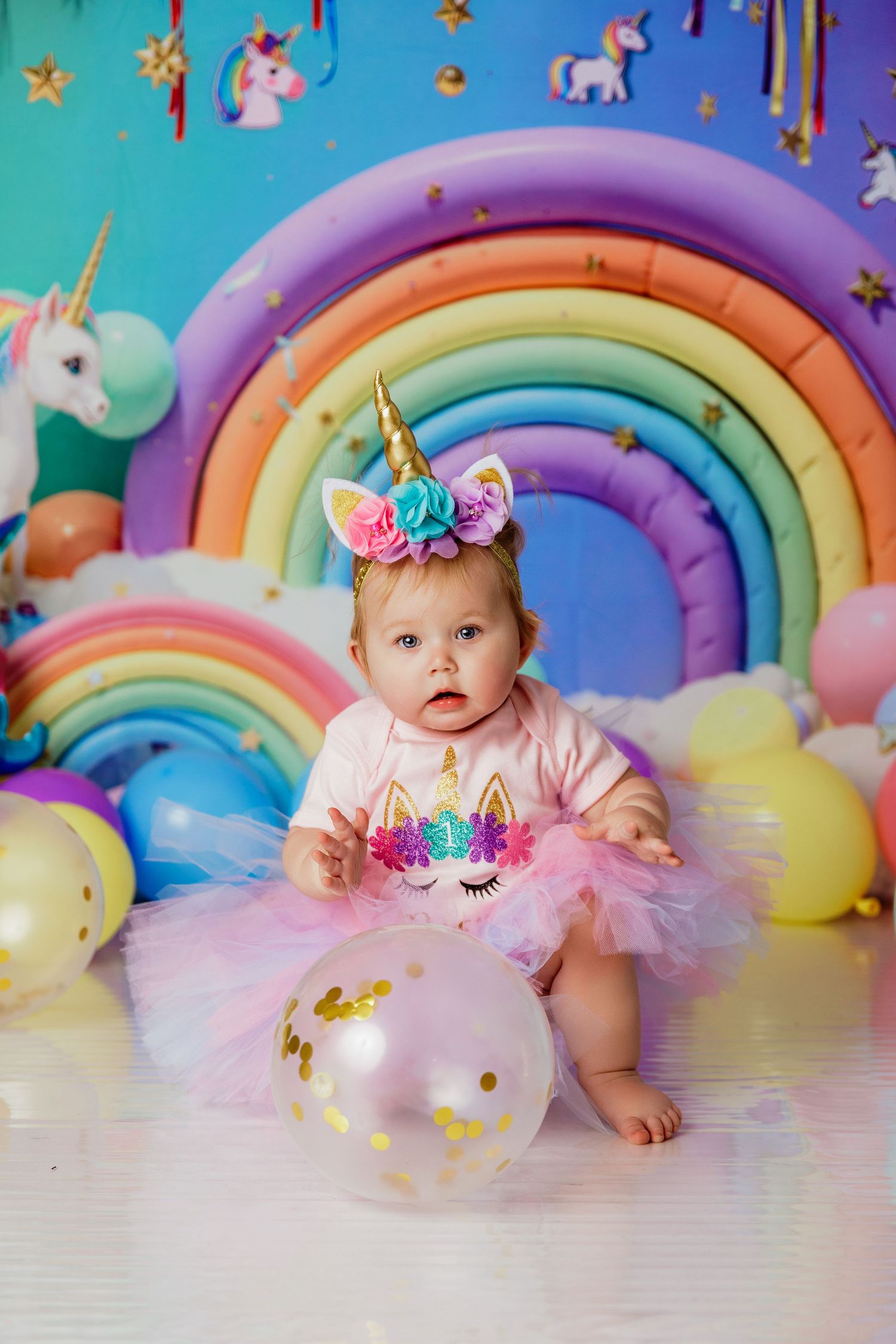 Baby girl in unicorn headband and pink tutu sits among colorful balloons and rainbow backdrop at birthday party.