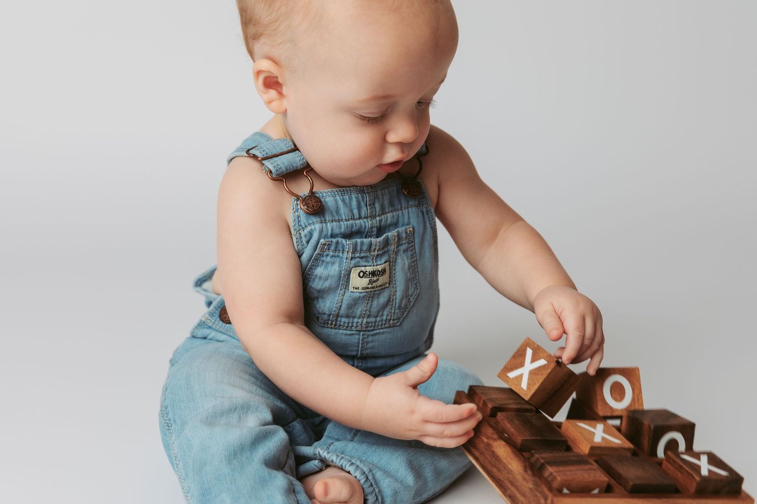 Baby in blue denim overalls plays with a brown leather bag while sitting on a light gray background.
