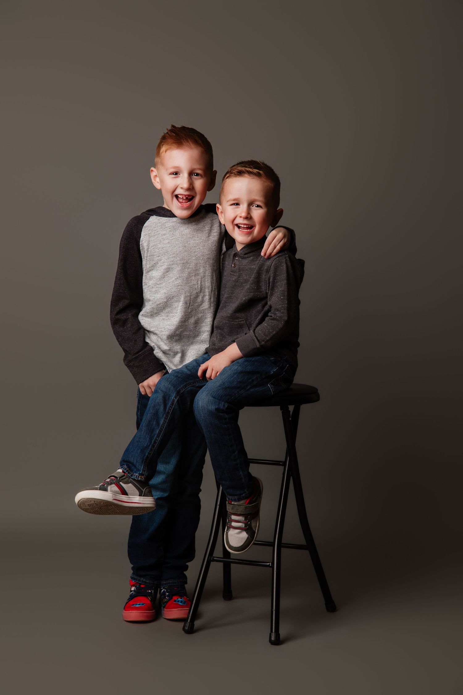 Two playful children laugh and interact while sitting on a black chair against a gray studio backdrop.