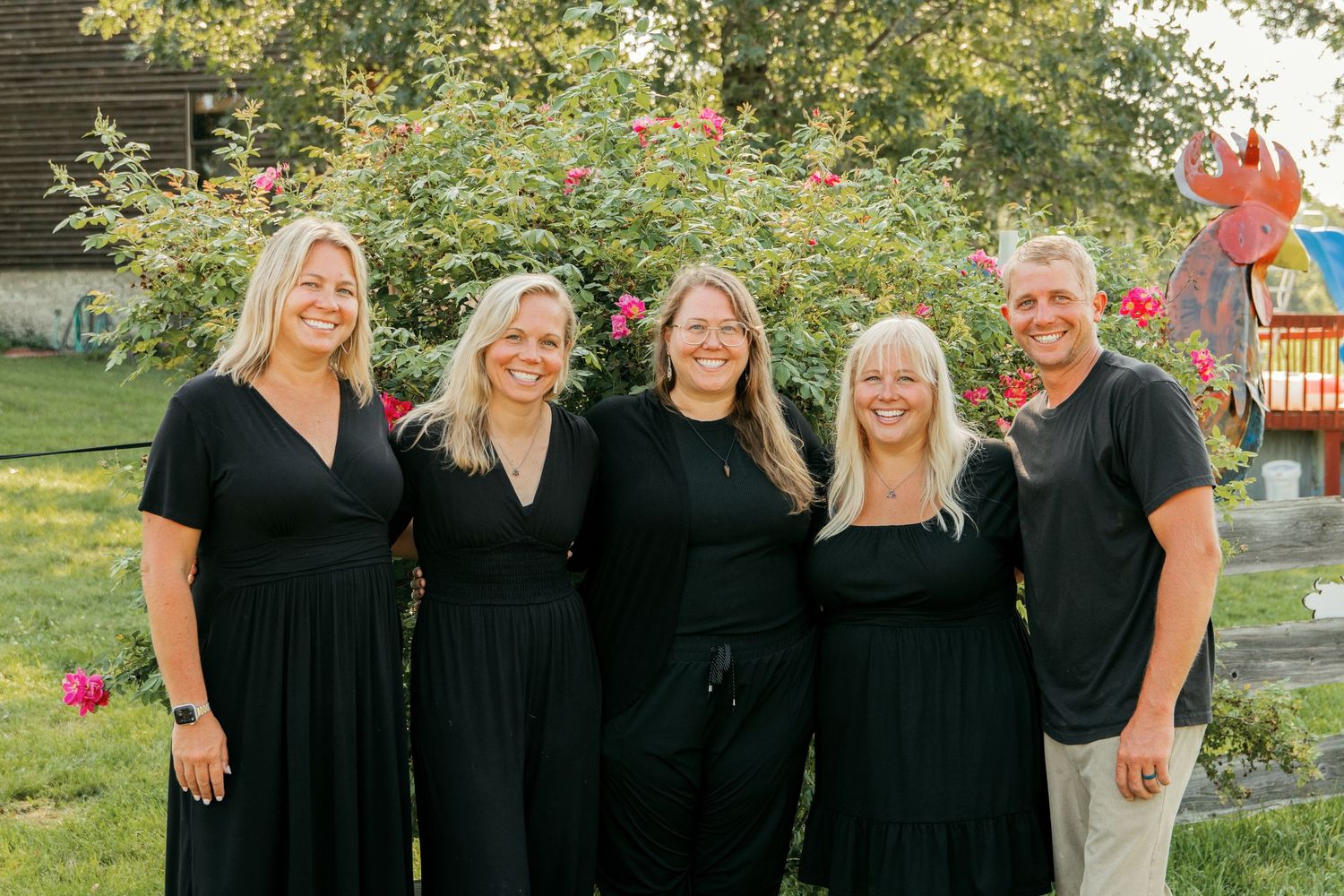 Group wearing matching black dresses pose together in garden with blooming roses behind them.