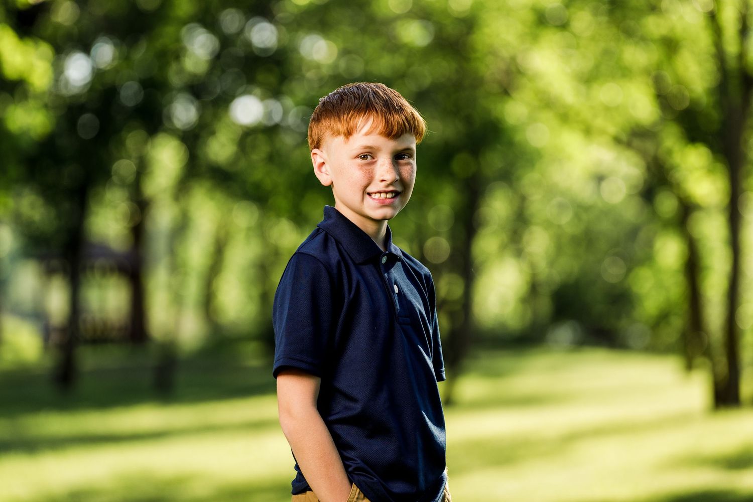 Smiling redhead boy in a navy polo shirt posing in a sunny green park for a portrait photo.
