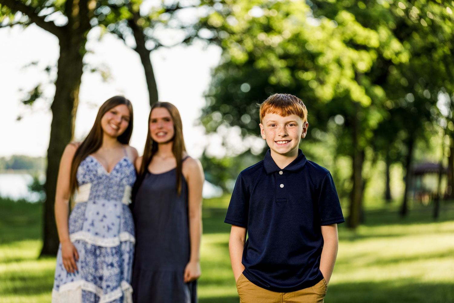 Young boy smiling in foreground with two girls standing behind him under green trees in a park.