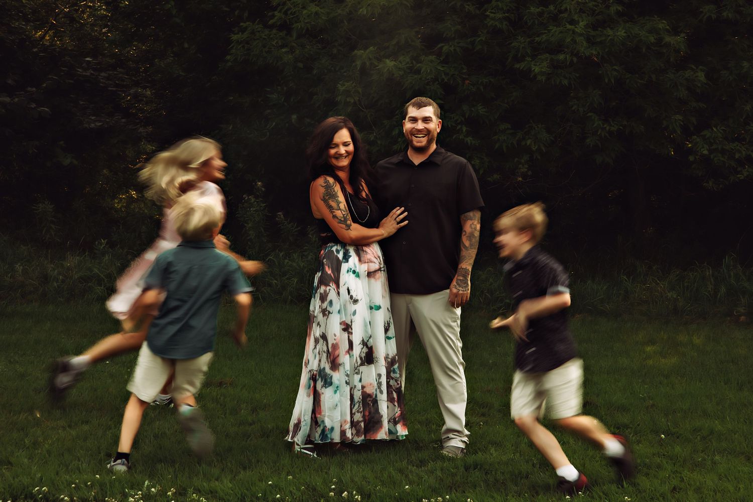 A couple smiles together outdoors while two children run past them in a lush green park setting.