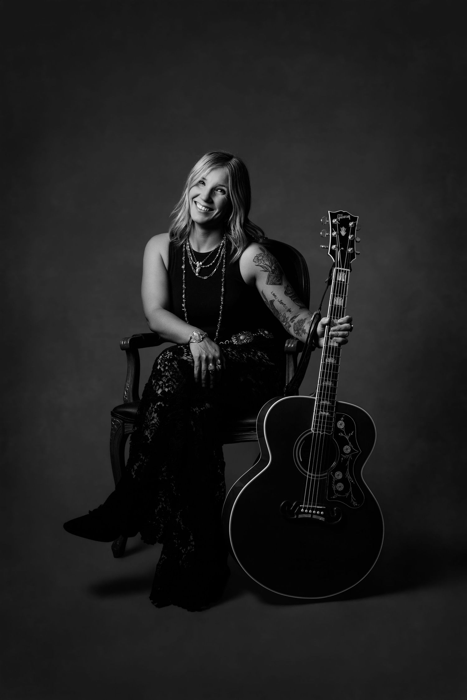 Black and white photo of a blonde female guitarist sitting in a chair holding an acoustic guitar, wearing laced pants.