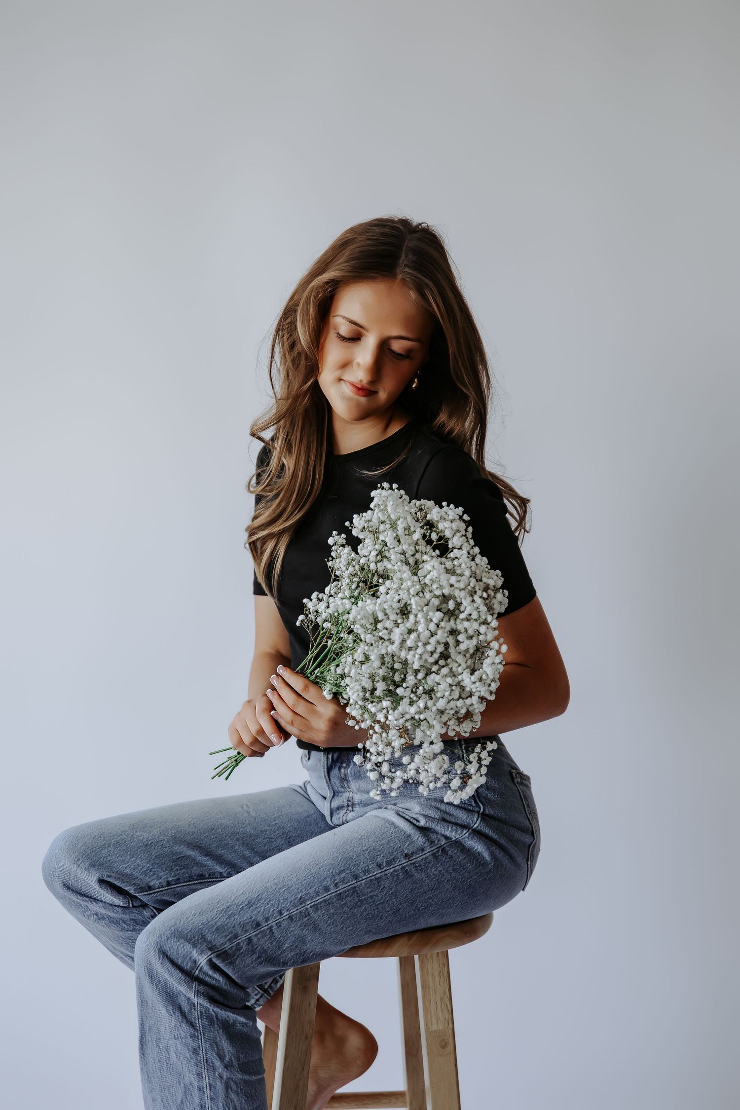 Young woman in black tee and jeans sits on wooden stool holding large bouquet of white baby's breath flowers.