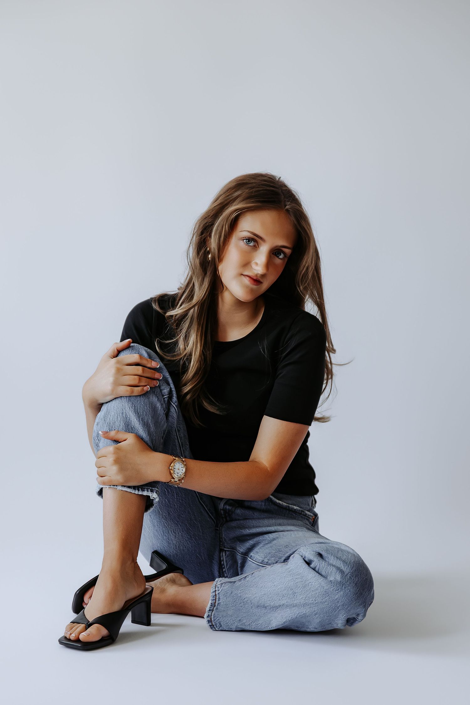 Young woman in black t-shirt and jeans sitting on floor hugging knee wearing black heeled sandals and a gold watch.