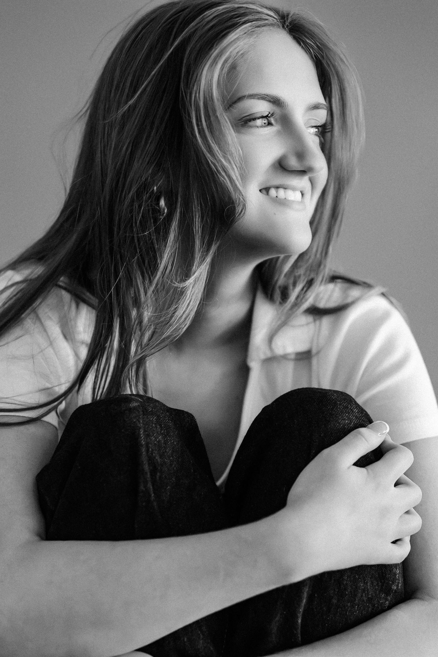 Black and white portrait of smiling young woman with flowing hair wearing white shirt hugging her knees.