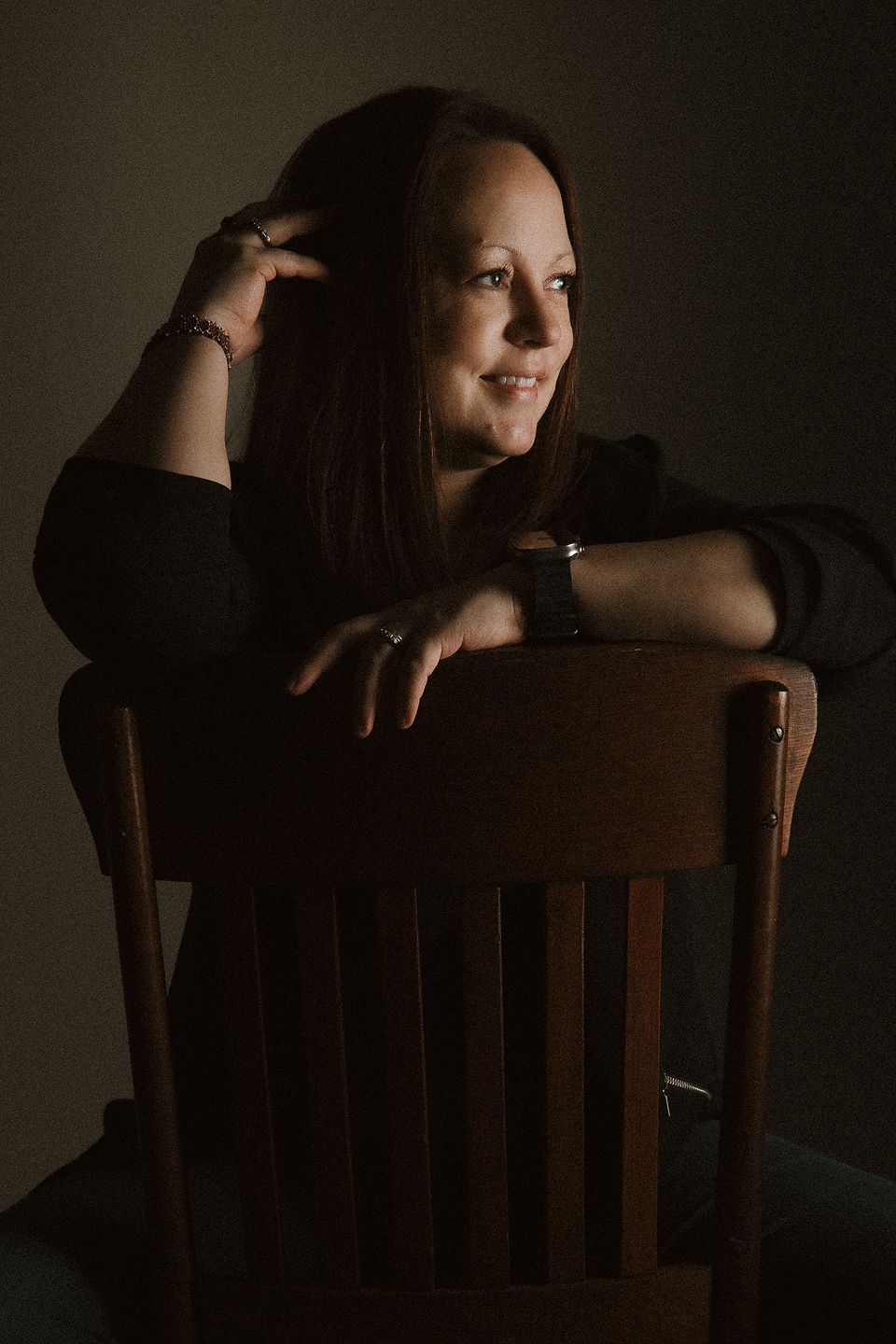 Woman in black top smiling, leaning on wooden chair back in dramatic low-key lighting portrait.