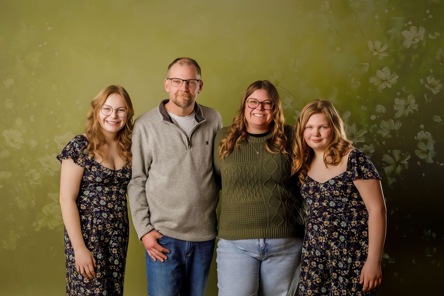 Family of four smiling together against a green floral backdrop, dressed in casual coordinated outfits.