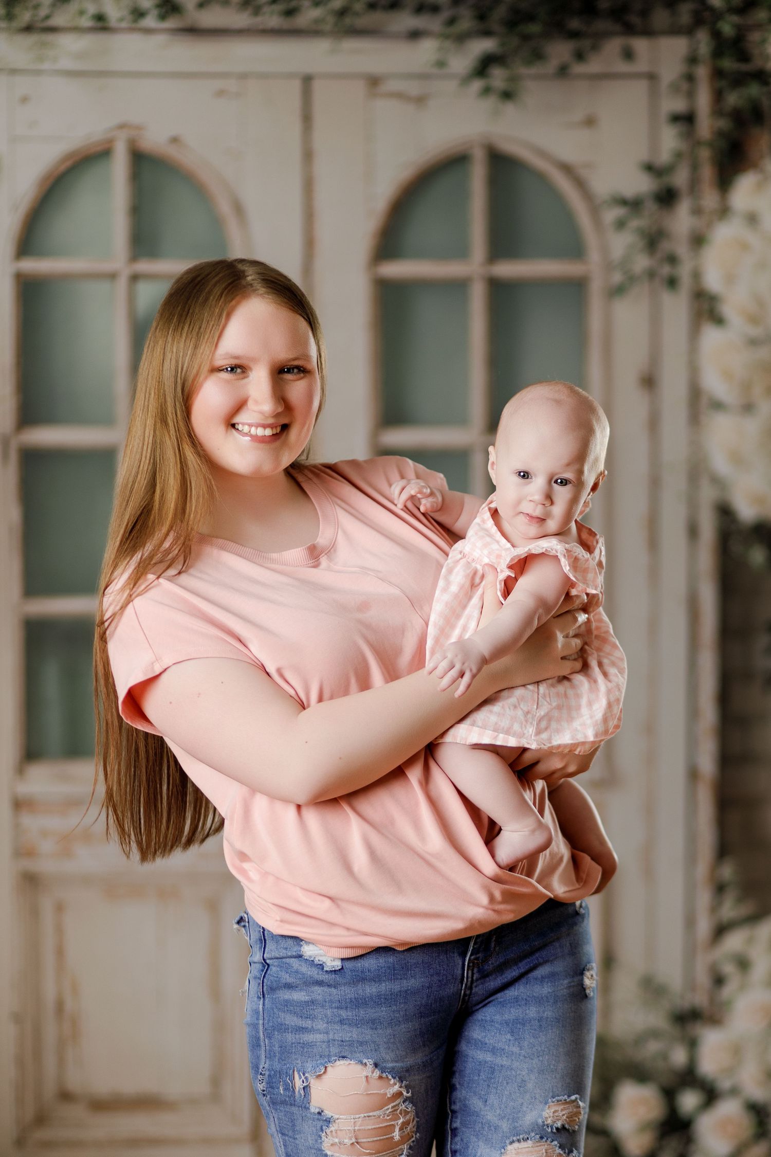 Smiling young woman in pink top holding a baby girl in a decorated studio with floral backdrop.