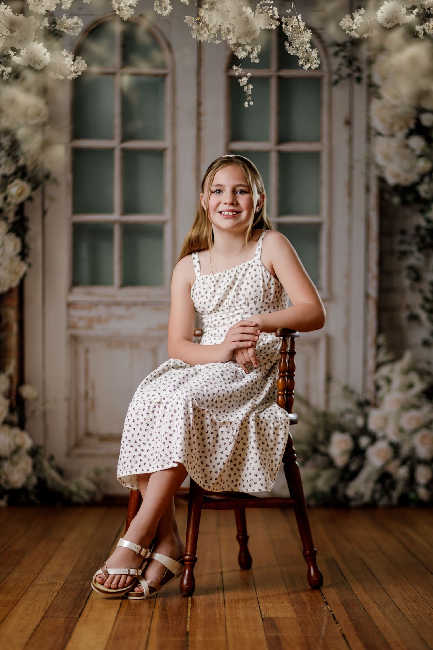 Young girl in polka dot dress sitting on wooden chair with floral backdrop.