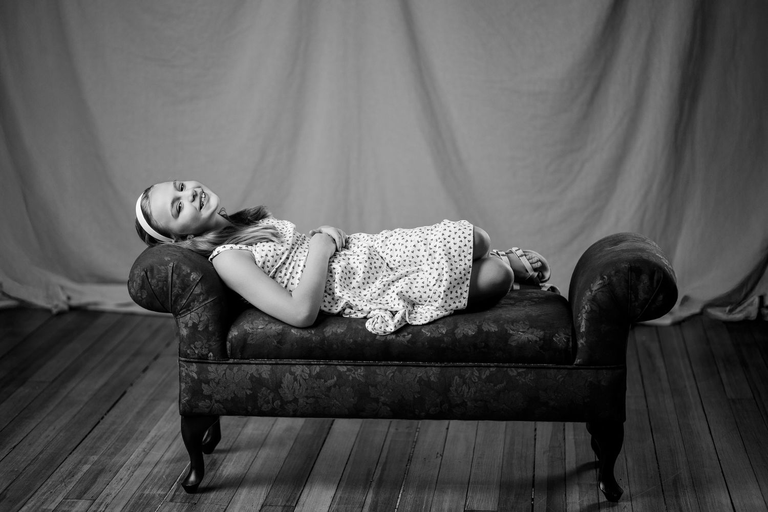 A young girl reclines on a vintage chaise lounge in a black and white portrait.