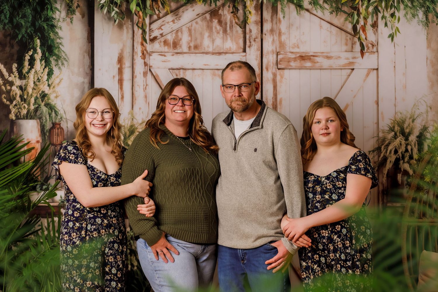 Family of four smiling together in front of rustic barn doors surrounded by lush greenery in a professional portrait.