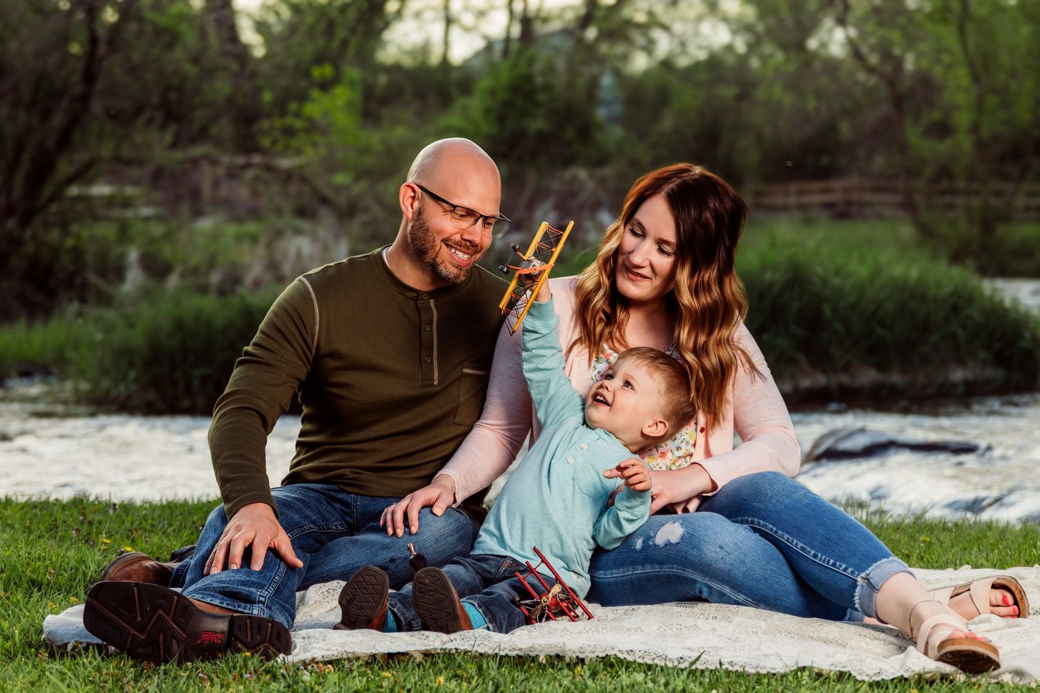 Happy family with parents and baby sitting on blanket in green park near river for outdoor portrait.