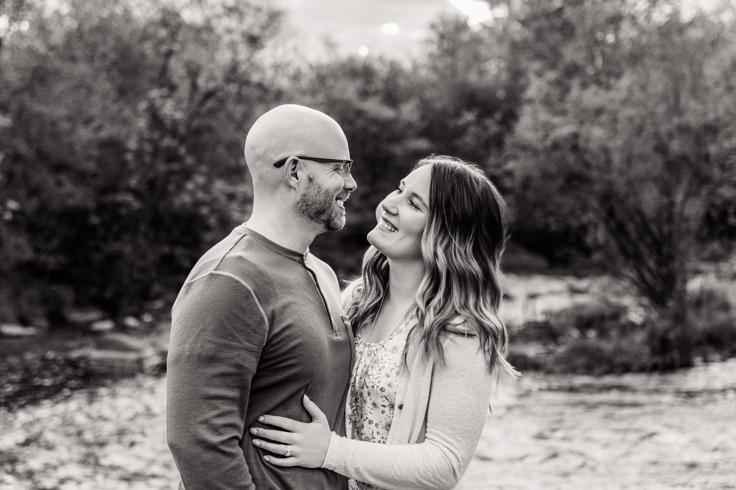 Black and white photo of a smiling couple embracing outdoors among trees during an engagement session.
