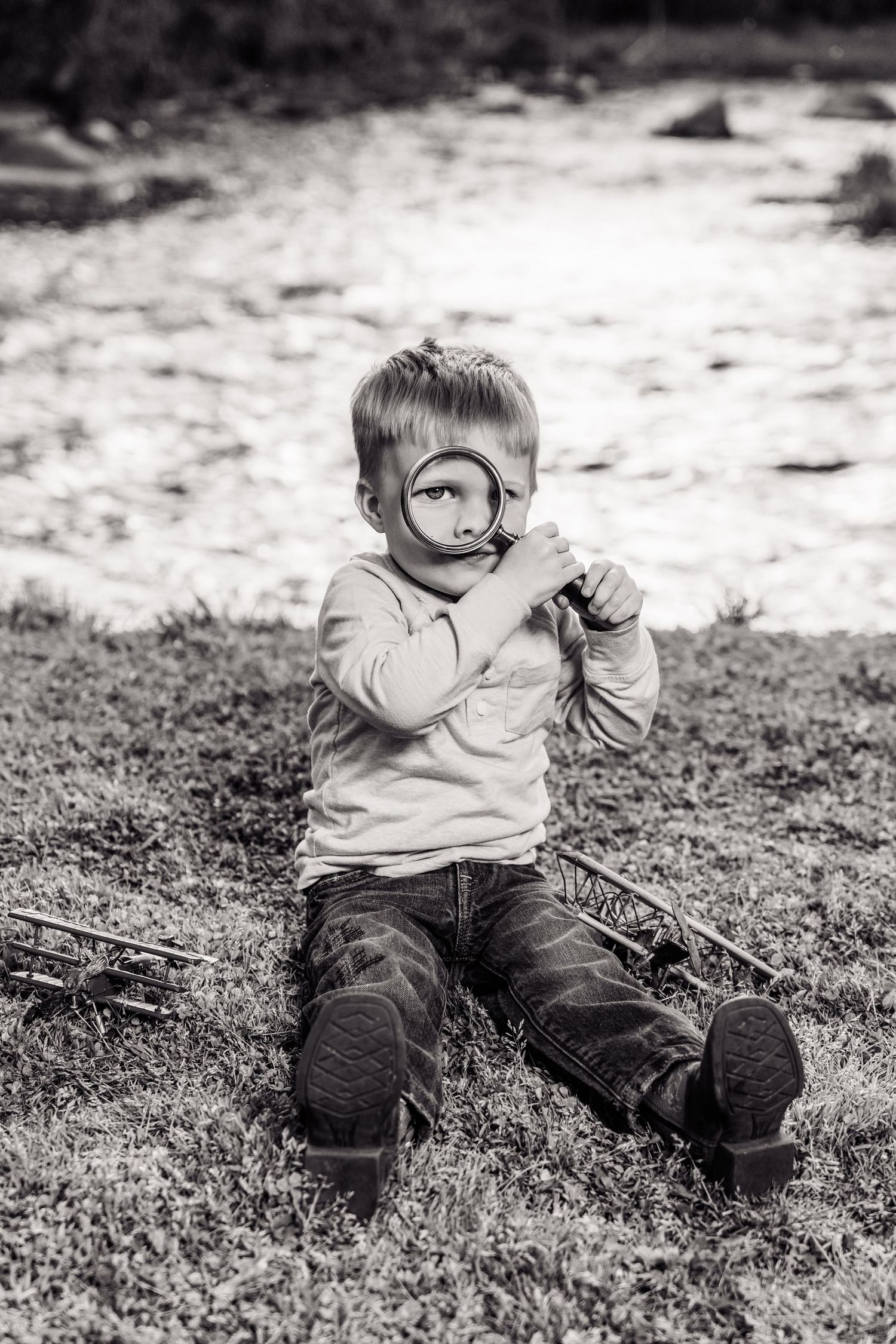 Black and white photo of a young boy sitting on grass, holding a magnifying glass up to his eye outdoors.