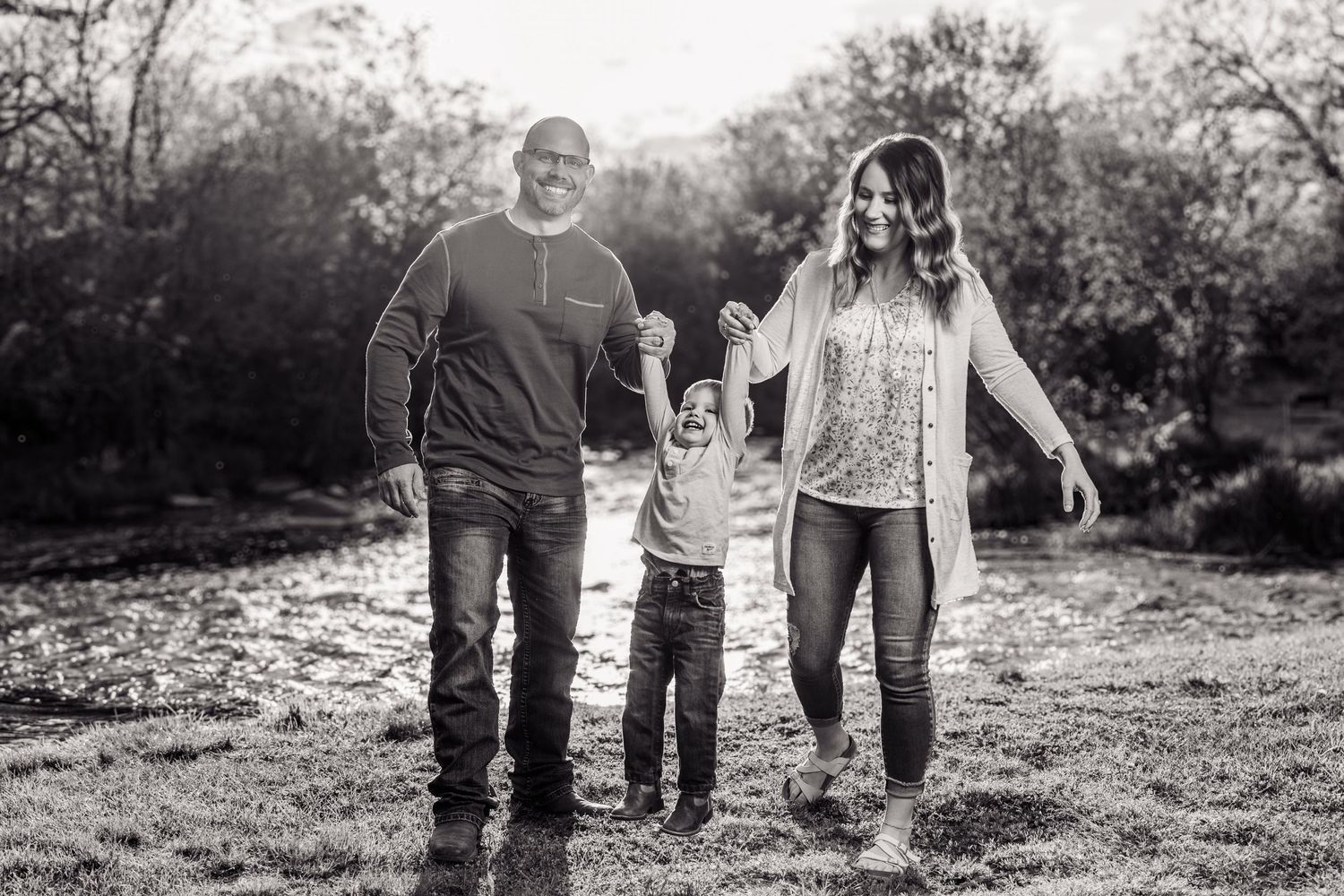 Black and white family portrait of parents holding hands with young child outdoors in natural setting.