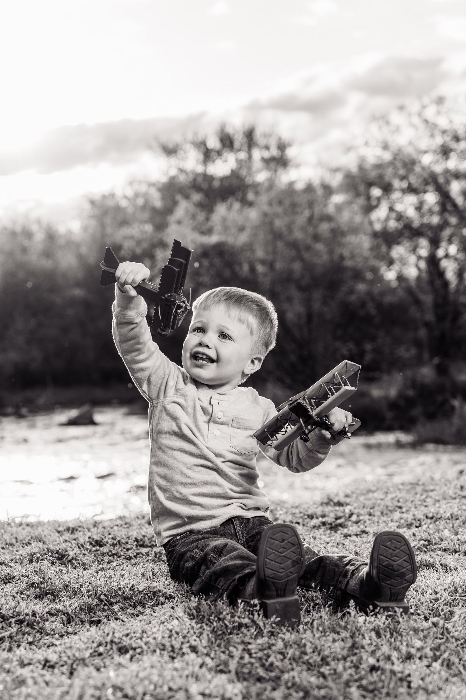 Black and white photo of a happy toddler sitting on grass, joyfully holding toy airplanes up in the air outdoors.