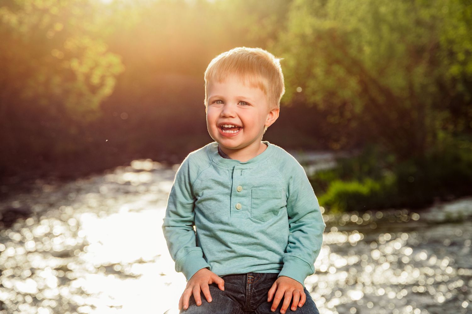 Happy toddler boy laughing outdoors near a sunlit river, wearing a teal shirt and jeans on a sunny day.