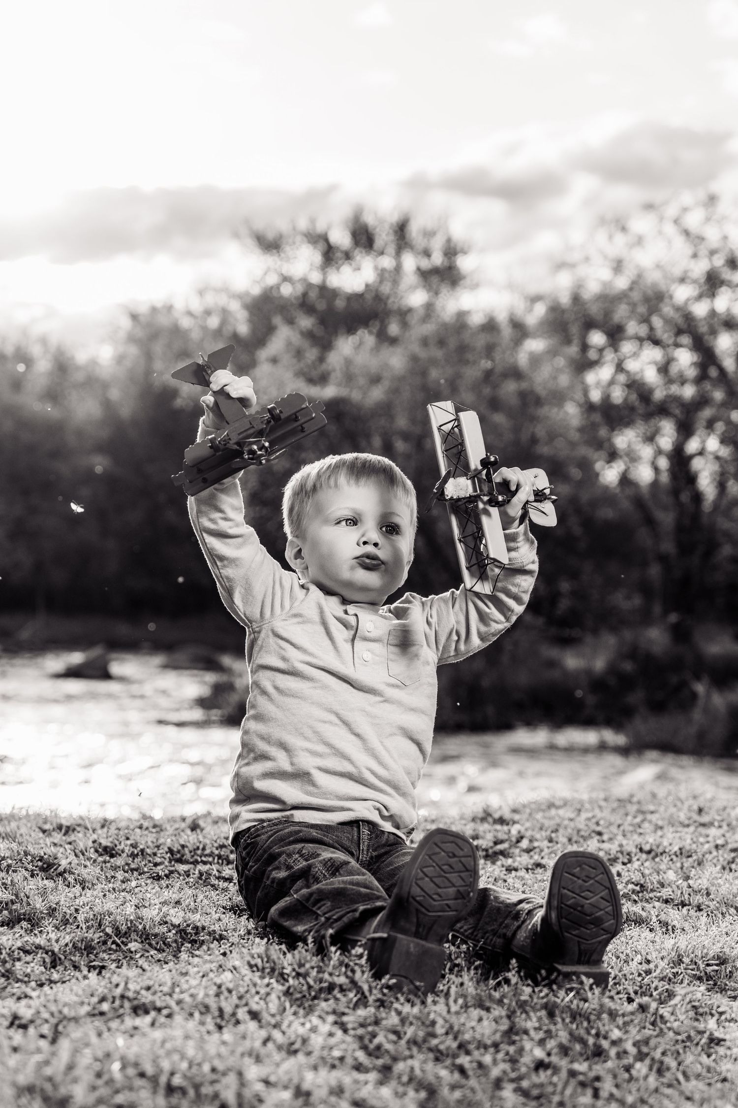 Black and white photo of a toddler boy sitting on grass, joyfully holding up two toy planes outdoors.